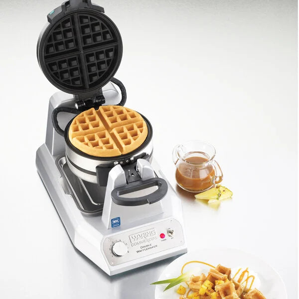 Waring waffle maker with a waffle inside, accompanied by a small bowl of syrup and a plate of waffles on a white background.